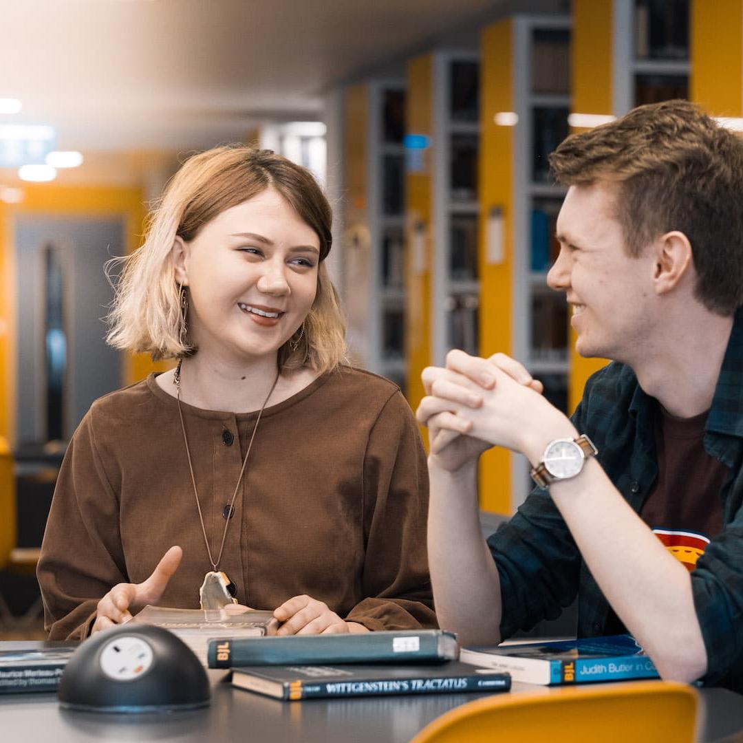 Philosophy students, Ellie Palmer and Ben John Campion, sit smiling at a desk in the Brynmor Jones Library.