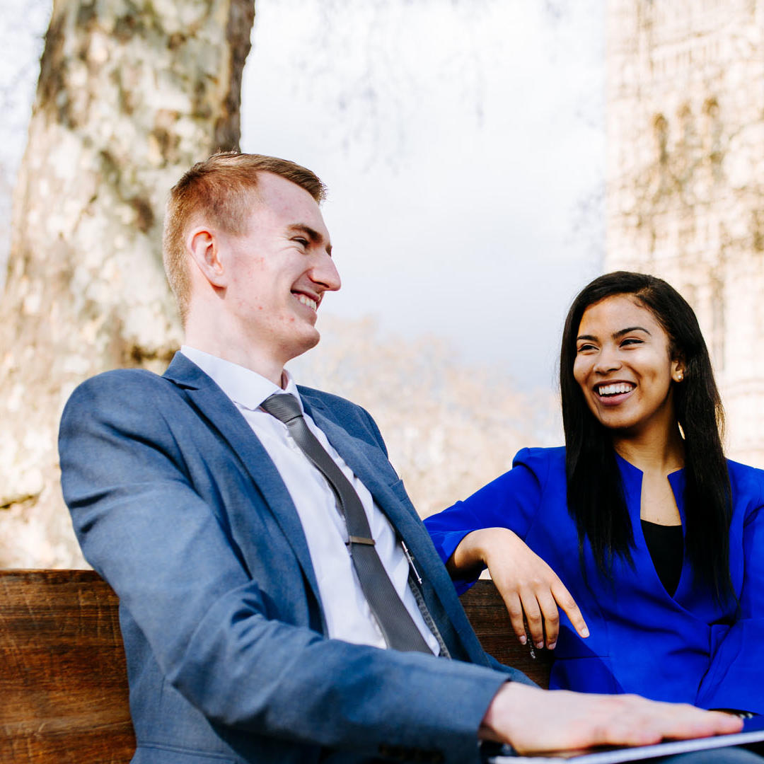 Two Hull Politics students sit smiling on a bench outside the Houses of Parliament while on placement in Westminster.