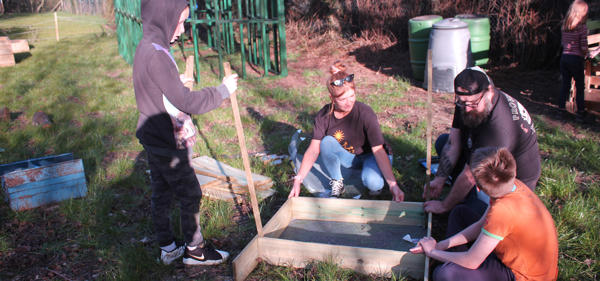A group of children are outdoors with two youth workers, creating a composting box out of wood